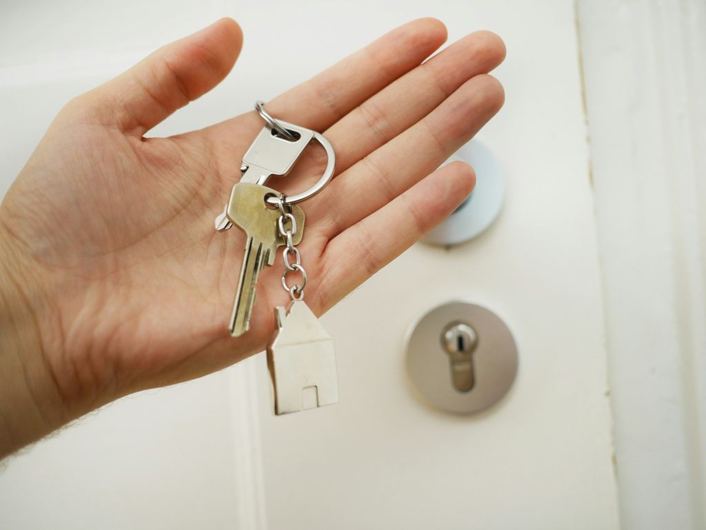 Geöffnete Hand mit einem Schlüsselbund vor einer weißen Wohnungstür, daneben ein Profilzylinder im Schutzbeschlag
